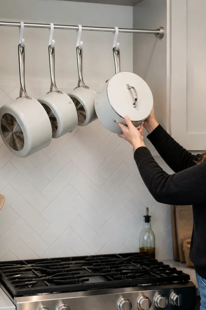 A tension rod spans above a small kitchen sink area. Pots hang from white enamel hooks, and a few white mugs hang beside them.